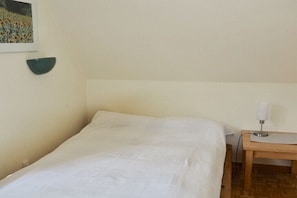 Ein Einzelbett mit weißer Bettdecke, ein kleiner Holztisch mit Lampe und eine Wandleuchte.