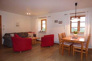 Ein Zimmer mit einer Couch, einem Esstisch mit Stühlen, einem Fenster mit Vorhängen und einer Heizung.