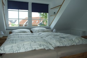 Ein Schlafzimmer mit großem Fenster, einem Bett mit gemusterten Betttextilien und einer schrägen Decke.