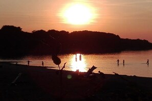 Ein Sonnenuntergang über einem Strand, mit stilisierter Darstellung von Personen und Bäumen.