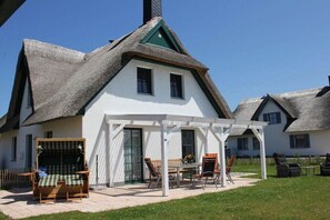 Ein weißes Haus mit einem Strohdach, einer überdachten Terrasse mit Stühlen und einem Tisch sowie ein kleiner Garten.