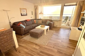 A living room with a sectional sofa, a small table, a wicker chair, and a view of the beach through large windows. - Apartment in Borkum with swimming pool (Borkum)