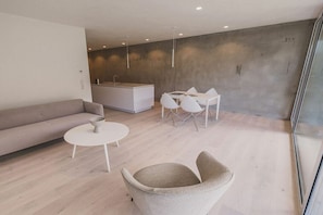 Ein modernes Wohnzimmer mit einer Couch, einem runden Couchtisch, einem beigen Sessel, einem Essbereich mit weißem Tisch und Stühlen sowie einer Küchenzeile mit weißer Arbeitsplatte und Schränken.