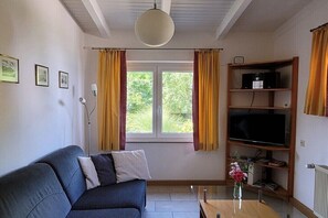 Ein Wohnzimmer mit einer Couch, einem Fernseher auf einem Holzschrank, einem Fenster mit Vorhängen und einer Vase mit Blumen auf dem Tisch.
