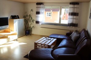 Ein Wohnzimmer mit einer Couch, einem Fernseher, einem Holztisch, einer Zimmerpflanze und einem Fenster mit Vorhängen.