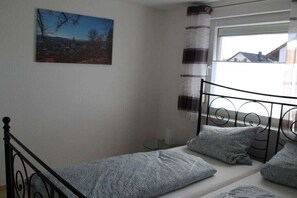 Ein Einzelbett mit Metallgestell, ein Fenster mit gestreiften Vorhängen und ein Landschaftsbild an der Wand.