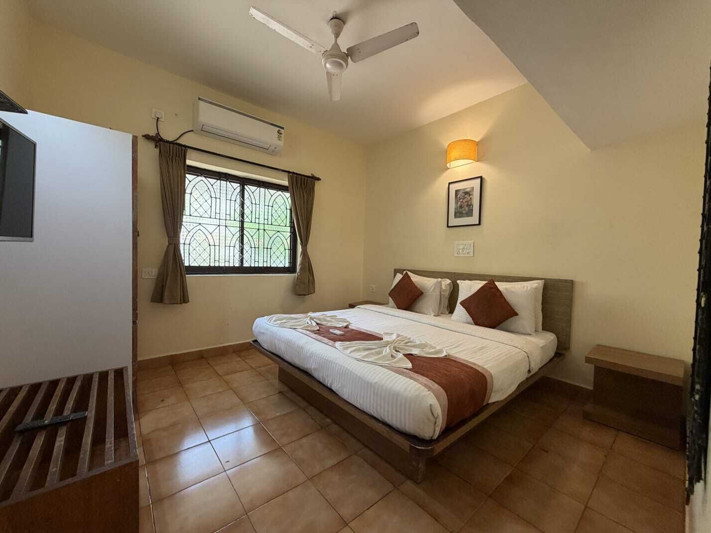 Een slaapkamer met een bed, een plafondventilator, een airconditioner, een raam met gordijnen, een nachtkastje en een wandlamp.