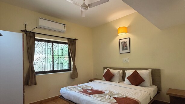 A bedroom with a bed, a ceiling fan, an air conditioning unit, a window with curtains, and a bedside table. - Lifestyle Villa (Candolim)