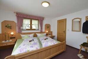 Ein Doppelbett mit floralem Bettzeug, ein hölzernes Kopfteil, Nachttischlampen, ein Fenster mit Vorhängen, eine Holztür, ein Spiegel und ein kleiner Tisch mit Schuhen.