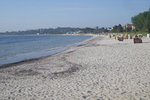 Una playa con arena, un mar tranquilo y una costa lejana con edificios y árboles.