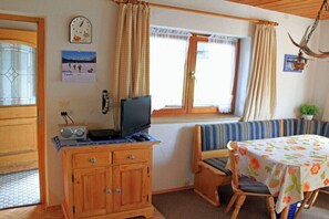 Ein Zimmer mit einem Holzschrank, einem Fernseher, einer Bank, einem Tisch mit blumigem Tischläufer und einem Fenster mit Vorhängen.