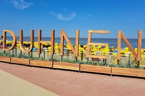 Ein hölzernes Schild mit dem Wort 'DUHENEN' in großen Buchstaben, ein Strand mit gelben Sonnenschirmen und einem wolkenlosen, blauen Himmel.