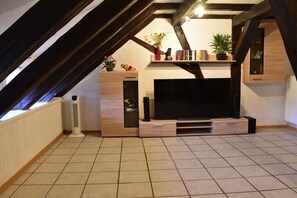 Ein Zimmer mit einem großen Flachbildfernseher, Holzbalken, einem Regal mit Büchern und Pflanzen sowie einer Heizung.