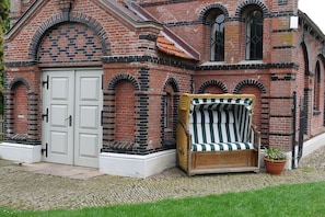 Ein Backsteinhaus mit weißer Tür und Fenstern, eine Bank mit gestreiften Kissen und einem Blumentopf.