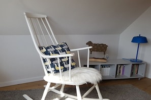 Ein weißer Schaukelstuhl mit einem flauschigen Sitz und einem gemusterten Kissen, ein Bücherregal mit Büchern, eine blaue Lampe und eine Holz-Schaf-Dekoration.
