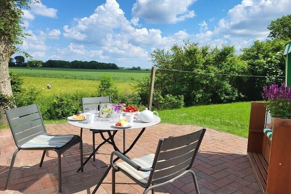 Eine Terrasses mit Tisch für zwei Personen, umgeben von Stühlen, mit Blick auf ein grünes Feld und Bäume.