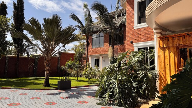 Una casa de ladrillo con balcón, un patio empedrado con baldosas estampadas y diversas plantas tropicales.
