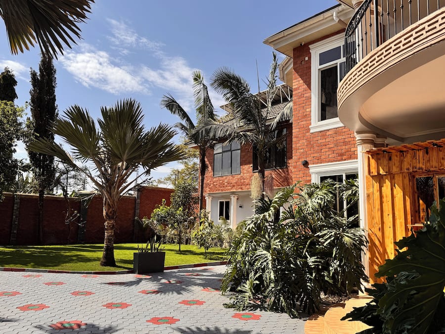 A brick house with a balcony, a paved courtyard with patterned tiles, and various tropical plants.