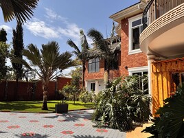A brick house with a balcony, a paved courtyard with patterned tiles, and various tropical plants.