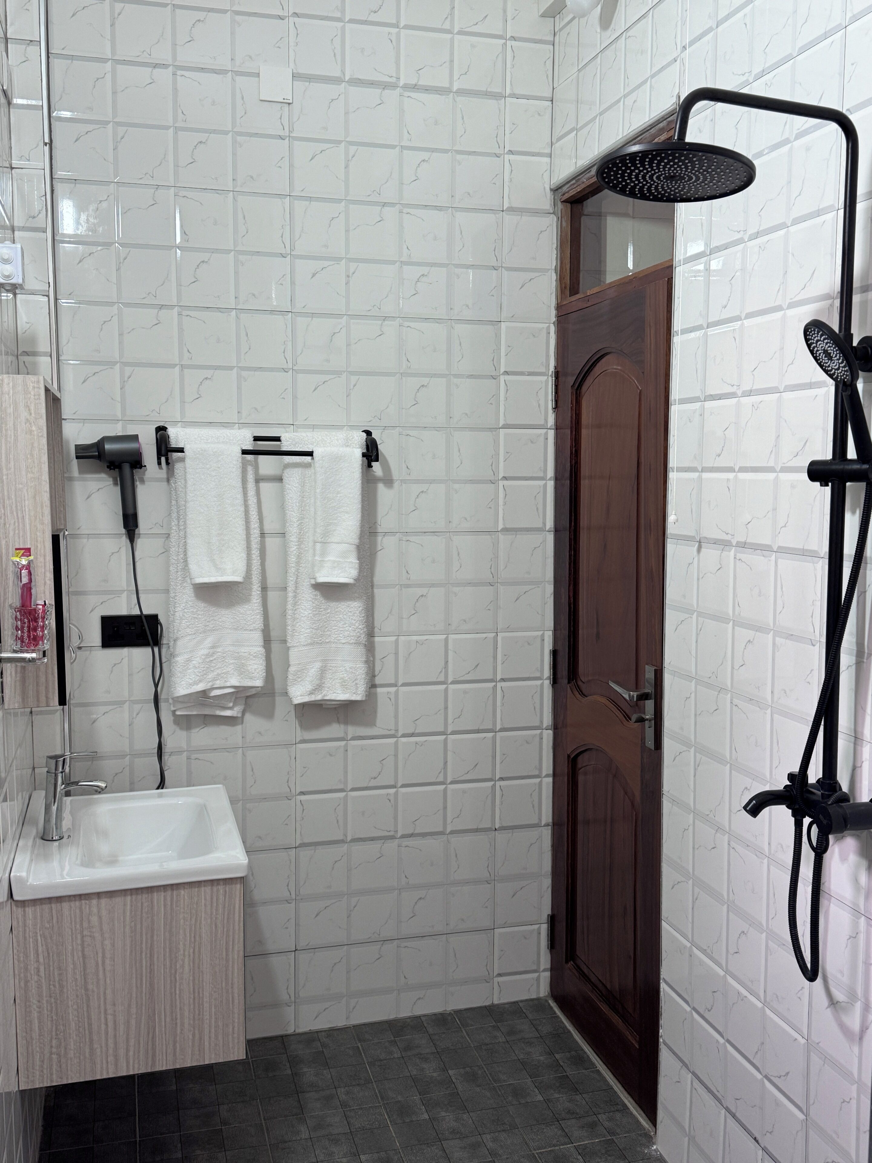 A bathroom with white marble tiles, a wooden door, a white sink, and a black showerhead.