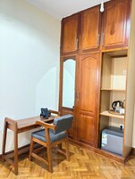 A room with a wooden desk, a chair, a wardrobe, and a small kitchenette.