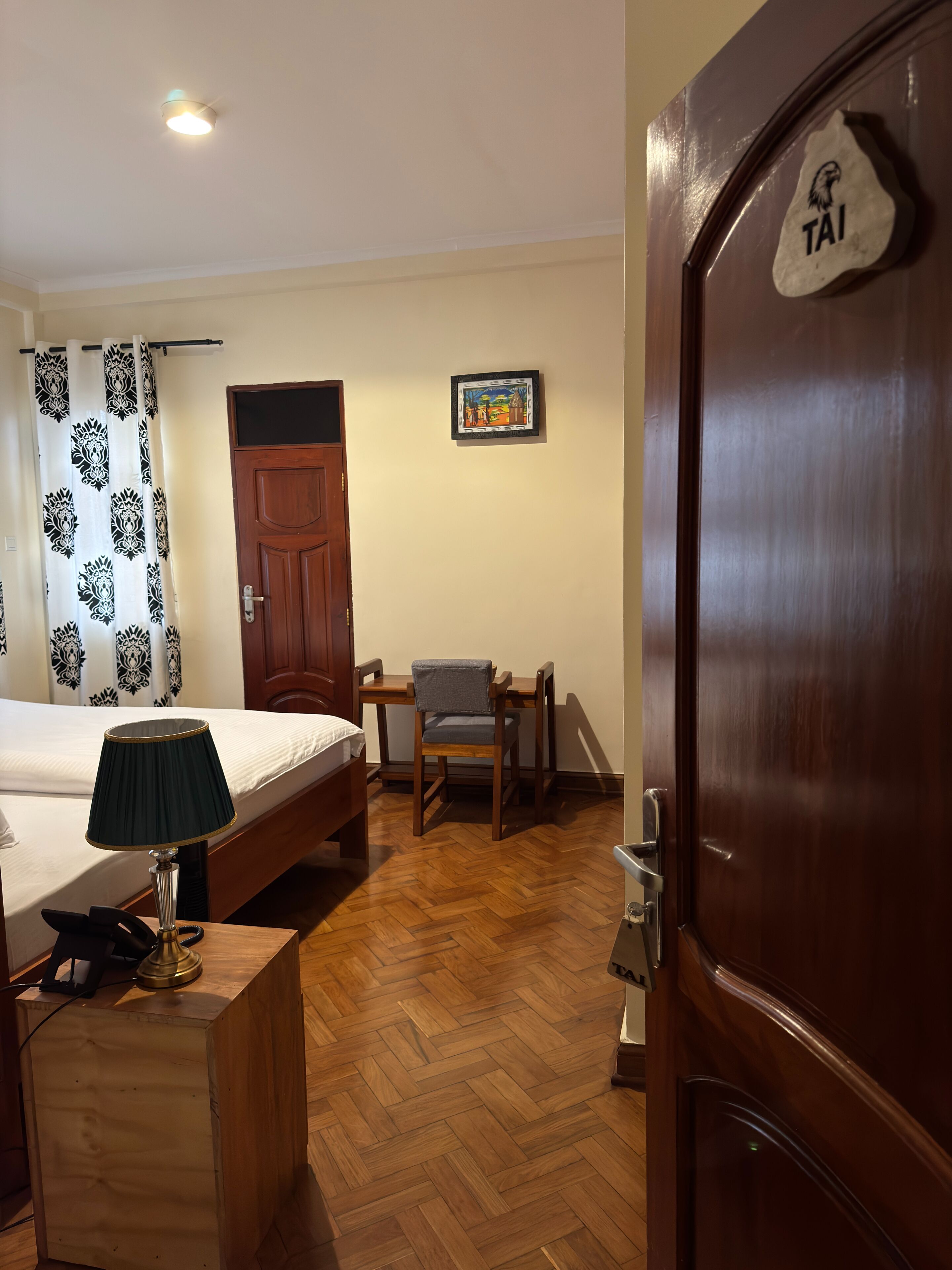 A hotel room with a bed, a desk with a lamp, a chair, a wooden floor, a door with a 'TAI' sign, and a framed picture on the wall.
