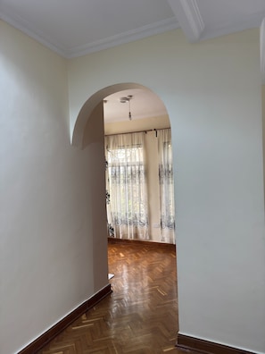 Un couloir doté d’un parquet en bois disposé en chevrons, de murs blancs et d’une grande porte en arc donnant sur une pièce avec des rideaux à motifs et un luminaire au plafond.