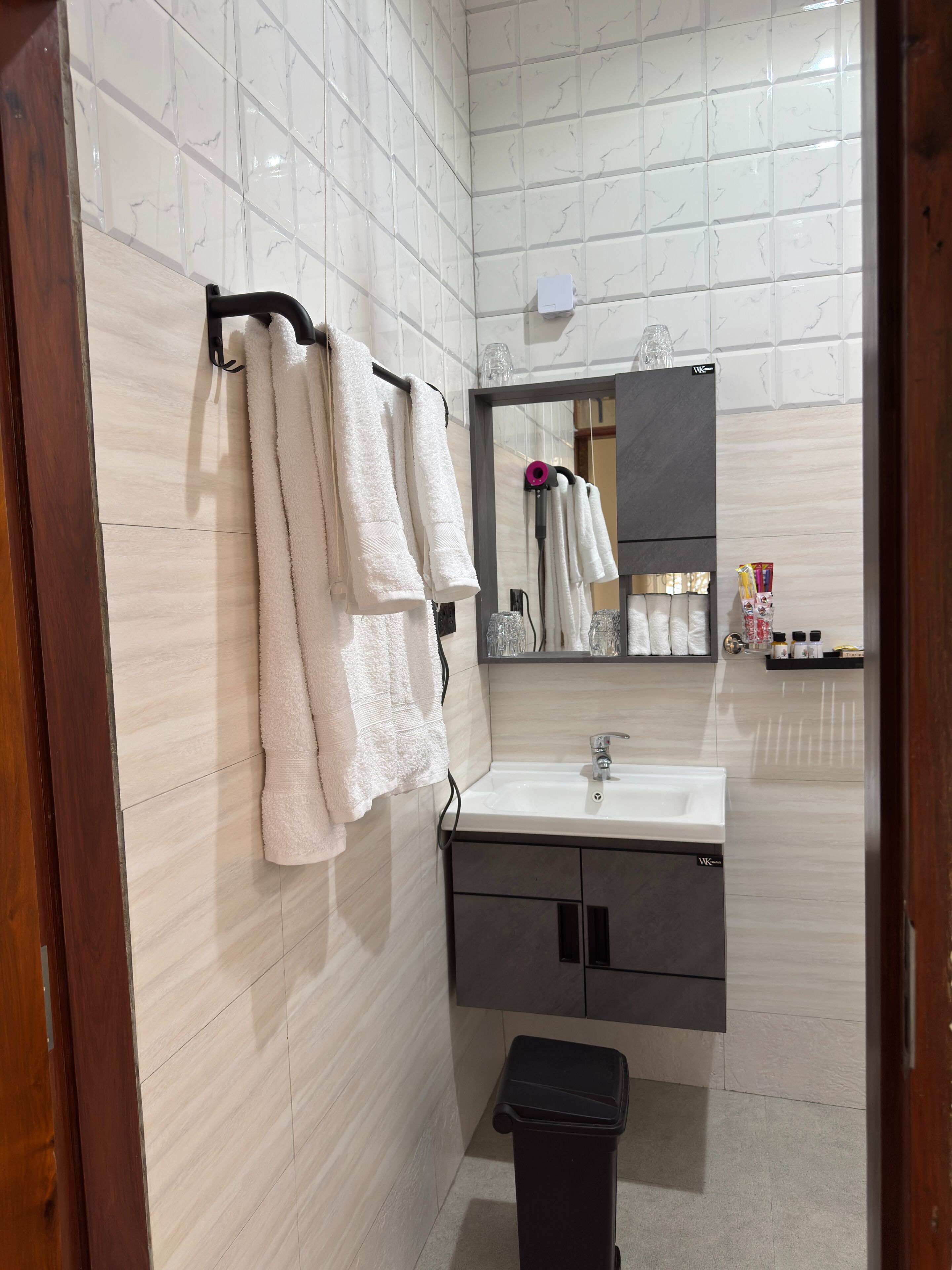 A bathroom with white tiled walls, a sink, a mirror, and a black trash bin.