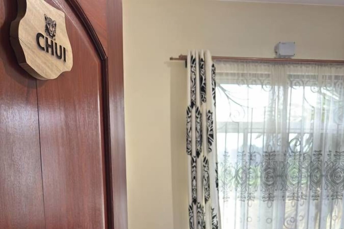 Una puerta de madera con un letrero, una cama con ropa blanca, una ventana con cortinas estampadas y una mesita con una lámpara.