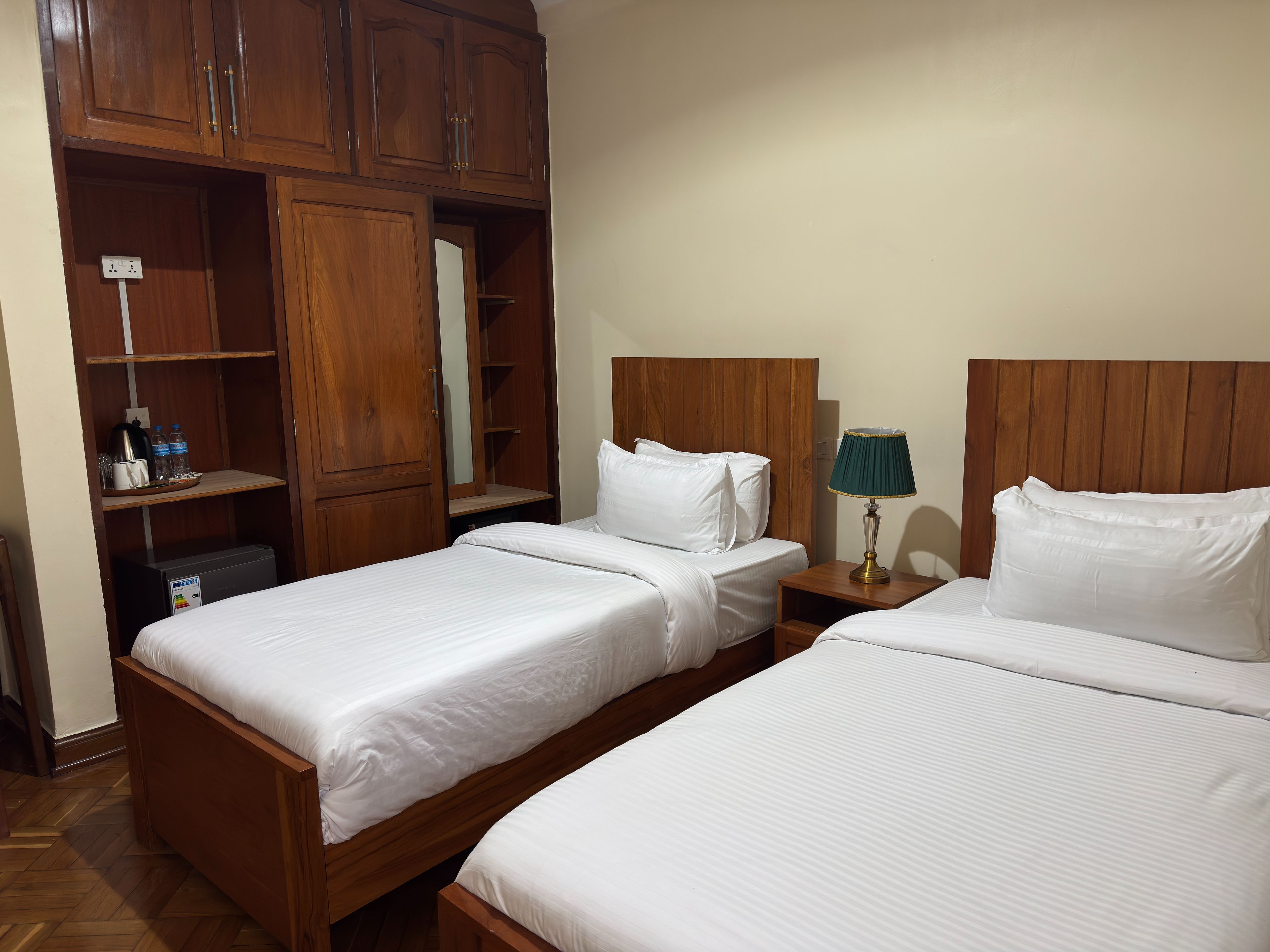 Une chambre d’hôtel avec deux lits simples, des têtes de lit en bois, une table de chevet avec une lampe et une armoire avec un miroir.