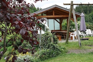 Eine Holzhütte mit Dach, umgeben von Grünpflanzen und weißer Gartenmöbeln.