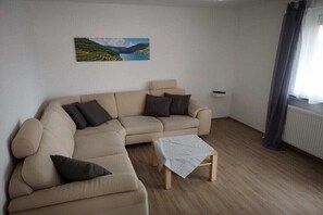 A living room with a beige sectional sofa, a small wooden table, and a painting of a landscape on the wall. - Holiday apartment in Werbach with barbecue (Werbach)