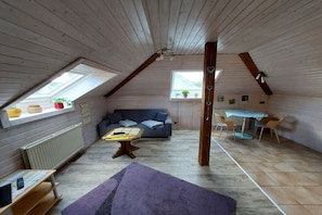 Ein gemütliches Dachzimmer mit schrägen Wänden, einer Couch, einem kleinen Tisch und einer Essecke mit Stühlen.