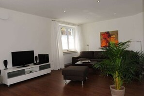 Ein modernes Wohnzimmer mit Flachbildfernseher, Lautsprechern, einer Couch, einem Hocker, einer Zimmerpflanze und einem Bild an der Wand.