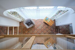 Ein Zimmer mit schrägen Wänden, einem Holztisch mit Zeitschriften, einem grauen Sofa und Blick durch ein Fenster mit Vorhängen.