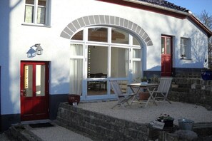 Ein weißes Gebäude mit roter Tür, einem großen Bogenfenster und einer kleinen Terrasse mit Tisch und Stühlen.
