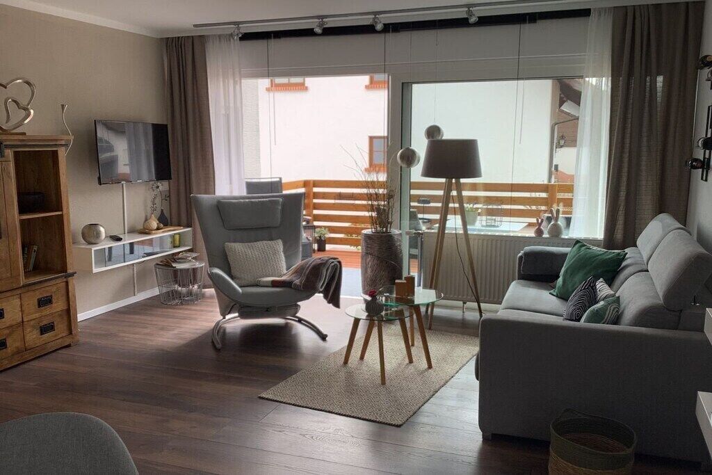 Ein Wohnzimmer mit grauem Sofa, grauem Sessel, einem kleinen Tisch mit Lampe, einem Fernseher auf einem Schrank und Blick auf den Außenbalkon.