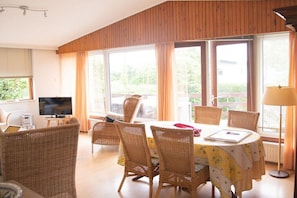 Ein Zimmer mit Esstisch, Korbstühlen, Fernseher und großen Fenstern.