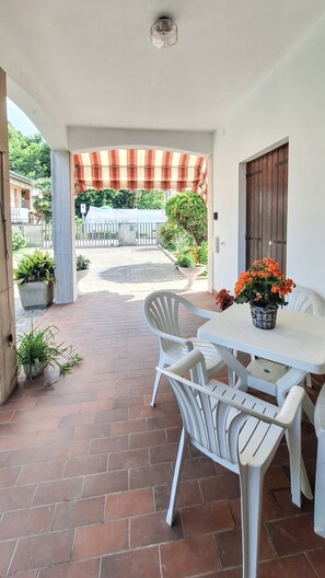 Eine überdachte Terrasse mit Tisch und Stühlen, Blumentöpfen und Blick auf einen Garten sowie Gebäude im Hintergrund.