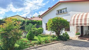 Ein weißes Gebäude mit rotem Dach, ein Garten mit verschiedenen Pflanzen und Blumen sowie ein überdachter Außenbereich mit Sitzgelegenheiten.