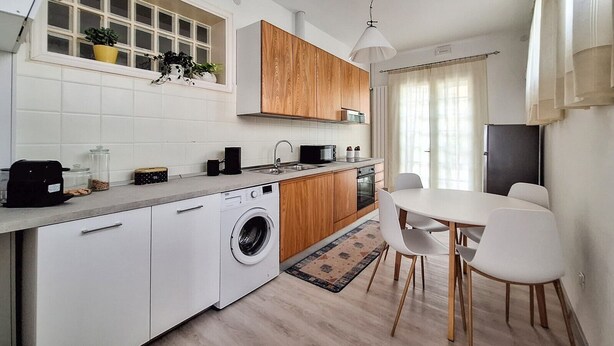 A modern kitchen with a built-in washing machine, wooden cabinets, a dining area with a table and chairs, and a window with plants. - Casa degli Orti in Venice (Dolo)