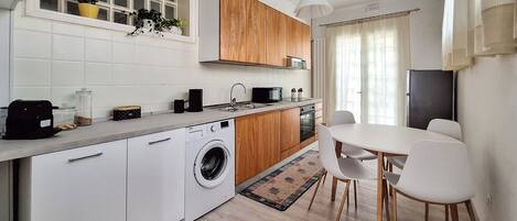 Eine moderne Küche mit Einbauwaschmaschine, Holzschränken, einer Essecke mit Tisch und Stühlen sowie einem Fenster mit Pflanzen.
