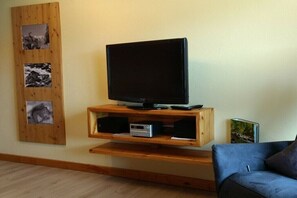 Ein Wohnzimmer mit einem Flachbildfernseher auf einem Holzboden, einem blauen Sofa und einem an der Wand befestigten Regal mit Bildern.