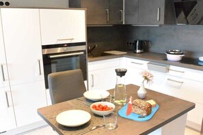 Eine moderne Küche mit weißen Schränken, einem eingebauten Ofen, einer Kaffeemaschine und einem Esstisch, gedeckt mit Tellern, einer Obstschale und einem Brot.