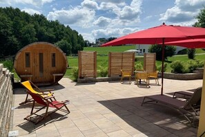 Eine Holzsauna, Sitzgelegenheiten im Freien und ein roter Sonnenschirm in einem Garten.