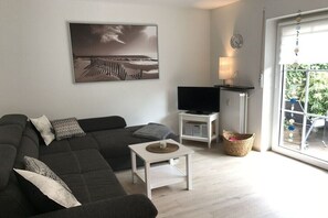 Ein Wohnzimmer mit einem dunkelgrauen Sofa, einem Flachbildfernseher, einem weißen Couchtisch und einem großen, gerahmten Bild an der Wand.