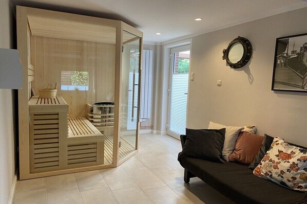 Ein Wohnzimmer mit einer Schiebetür zum Balkon, einem dunklen Sofa mit floralen Kissen und einem gerahmten Bild an der Wand.