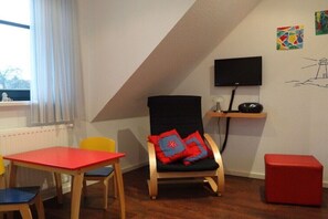 Ein Zimmer mit schrägen Wänden, einem an der Wand montierten Fernseher, einem roten Tisch, einem schwarzen Stuhl mit roten Kissen und einem kleinen roten Hocker.