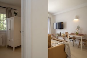 Ein Zimmer mit einem Bett, einem Fernseher, einem Esstisch mit Stühlen, einem Schrank und einem Fenster mit Vorhängen.