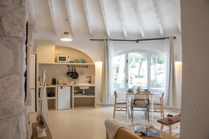 Une cuisine compacte équipée d’un four à micro-ondes, d’un évier et de divers ustensiles. On aperçoit un coin repas avec une table et des chaises, et une fenêtre offre une vue sur la verdure environnante.